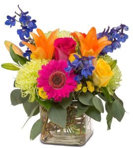 a variety of blossoms in vibrant colors. Roses, lilies, delphimium, mums and more arranged together to create this beautiful and colorful bouquet presented in a glass cube vase. 